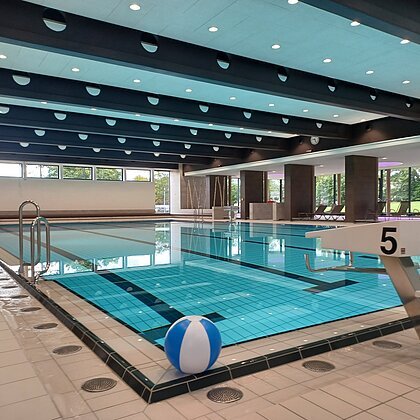 Innenaufnahme des leeren Schwimmbads - Jurabad Monheim. Zu sehen ist ein Schwimmbecken gefüllt mit strahlendem Wasser, ein Sprungbrett und ein weiß-blauer Wasserball liegt dekorativ im Vordergrund. Im Hintergrund des Hallenbads sind große Fenster zu sehen.