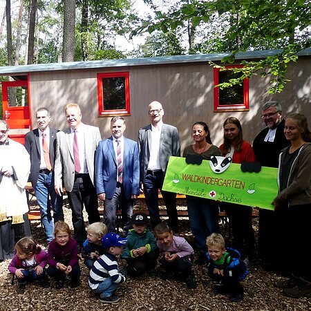 Eine Gruppe von Erwachsenen und Kindern steht bzw. kniet vor einem Bauwagen im Wald, drei Personen halten ein gestaltetes Schild mit der Aufschrift "Waldkindergarten Mandele Dachse" in den Händen.