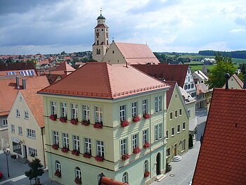 Haus des Gastes - im Hintergrund die Stadtpfarrkirche Monheim Blick von oben auf eine Innenstadt mit Hauptaugenmerk auf ein historisches grünes Gebäude im Vordergrund, mit roten Dachziegeln und Blumen an den Fenstern. Im Hintergrund weitere Häuser der Kleinstadt und blauer Himmel. Rechts unten im Eck das rote Dach eines anderen Gebäudes im Vordergrund zu sehen.