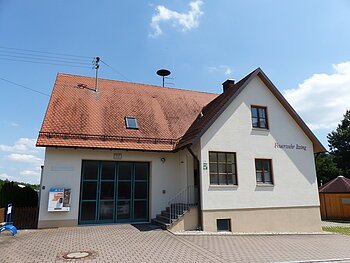 Ein Feuerwehrgebäude mit der Aufschrift "Feuerwehr Itzing" mit einem roten Ziegeldach, zwei Fenstern, einer Treppe und einem großen Tor. Im Hintergrund blauer Himmel und Bäume. Auf dem Gebäude ist ein Zigarettenautomat angebracht.