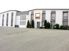 Weißes Gebäude mit mehreren Fenstern, Eingangsbereich und einem Parkplatz davor. Auf dem Gebäude in einem orangefarbenem Viereck der Schriftzug der Firma MKR. Vor dem Gebäude mehrere Thuja-Bäume und weitere Pflanzen.