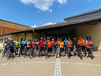 Gruppe von Menschen mit Fahrrädern und Helmen in bunter Kleidung vor einem Gebäude auf einem gepflasterten Platz.