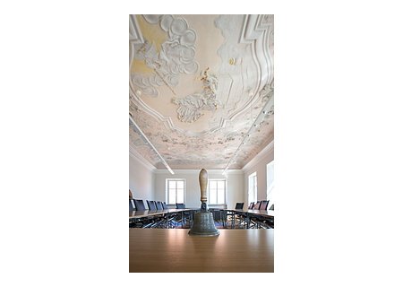 Im Vordergrund eine Handglocke mit Holzgriff auf einem Tisch in einem Sitzungssaal mit bemalter historischer Stuckdecke, die biblische Szenen zeigt und schwarze Stühle entlang des U-förmigen Tisches. Einige Fenster entlang der zwei Wände.