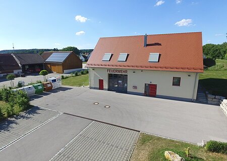 Gelbes Feuerwehrgebäude mit rotem Dach, Aufschrift "Feuerwehr" an der Fassade, zwei rote Türen, ein großes Tor und Fenster auf der Vorderseite. Vorderhalb großer gepflasterter Platz.  Im Hintergrund Häuser und blauer Himmel .