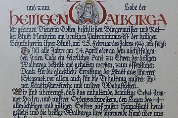 Ein handgeschriebenes Dokument mit dekorativen roten und schwarzen Buchstaben, das eine religiöse Widmung enthält. Hierin gelobt der Bürgermeister sowie der Rat der Stadt Monheim, ein alljährliches Dankamt zu Ehren der Heiligen Walburga abhalten zu lassen, für die Verschonung der Stadt im Zweiten Weltkrieg.