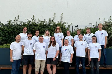 Gruppenfoto von 15 Personen in weißen T-Shirts mit dem Logo der Wasserwacht drauf, stehen blickend in die Kamera vor einer weißen Wand mit Pflanzen im Hintergrund.