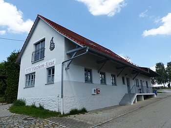 Weißes Gebäude mit rotem Dach, einer Treppe und einem Defibrillator. steht auf einem gepflasterten Grundstück. Auf dem Gebäude der Schriftzug "FFW Flotzheim-Kreut" an der Fassade. Blauer Himmel und Bäume im Hintergrund.