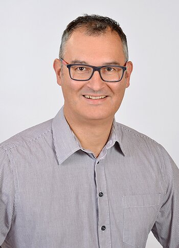 Porträt eines lächelnden Mannes mit Brille, grauem Hemd und kurz geschnittenem Haar vor weißem Hintergrund.