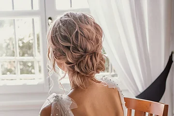 Frau mit hochgestecktem Haar und weißem, perlenbesetztem Kleid sitzt auf Holzstuhl vor Fenster mit weißen Vorhängen.