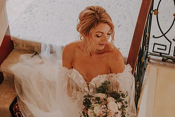 Braut in weißem Spitzenkleid mit Blumenstrauß steht auf Treppe und schaut nach unten.