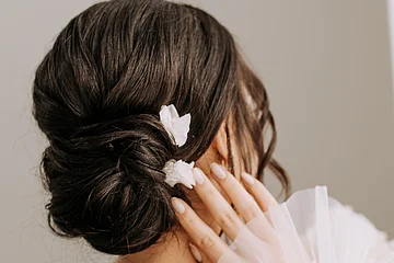 Frau mit hochgestecktem Haar und weißen Blüten, Hand berührt Gesicht, weißes Tüllkleid mit Perlendetails