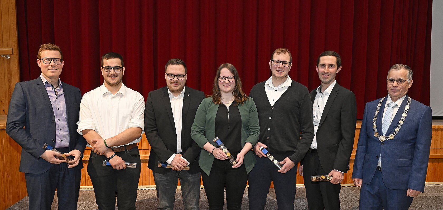 Sieben Personen stehen nebeneinander vor rotem Vorhang, sechs halten Auszeichnungen, eine trägt eine Amtskette. Ehrung am Neujahrsempfang 2026 der Stadt Monheim - Schützengesellschaft Monnheim mit dem Stadtmeister.