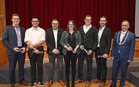 Sieben Personen stehen nebeneinander vor rotem Vorhang, sechs halten Auszeichnungen, eine trägt eine Amtskette. Ehrung am Neujahrsempfang 2026 der Stadt Monheim - Schützengesellschaft Monnheim mit dem Stadtmeister.