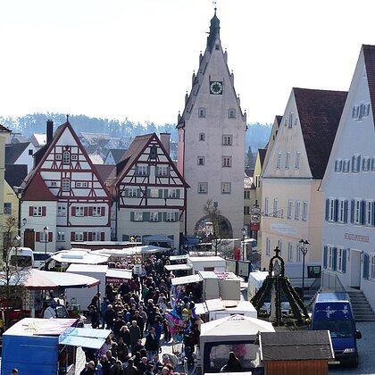 Josefsmarkt in Monheim in der Altstadt: auf dem Marktplatz entlang Fachwerkhäuser, ein Turm mit Uhr und Menschenmengen zwischen vielen Marktständen bei Tageslicht.