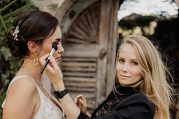 Frau trägt schwarzen Blazer und schminkt eine Braut in weißem Spitzenkleid vor Holztür draußen.