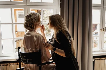 Frau trägt Seidenrobe sitzt auf Stuhl, während andere Frau ihr Make-up aufträgt vor großem Fenster.