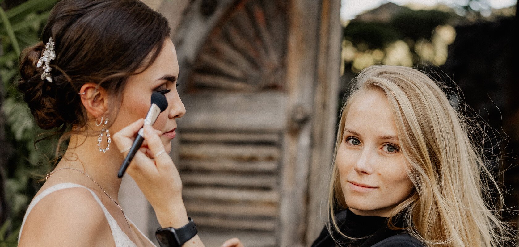 Frau trägt schwarzen Blazer und schminkt eine Braut in weißem Spitzenkleid vor Holztür draußen.