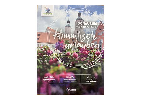 Blumen im Vordergrund, dahinter Gebäude mit zwei Türmen, Bayern-Logo, Text zu Donauries Ferienland Magazin 2026 "Himmlisch urlauben"