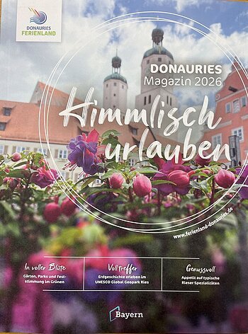 Blumen im Vordergrund, dahinter Gebäude mit zwei Türmen, Bayern-Logo, Text zu Donauries Ferienland Magazin 2026 "Himmlisch urlauben"