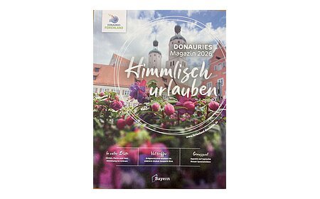 Blumen im Vordergrund, dahinter Gebäude mit zwei Türmen, Bayern-Logo, Text zu Donauries Ferienland Magazin 2026 "Himmlisch urlauben"