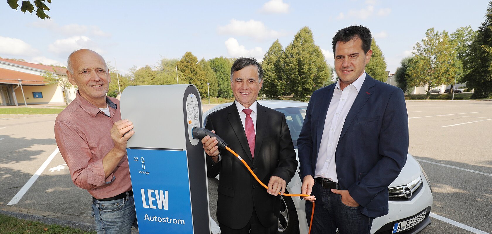 Drei Männer stehen neben einer LEW Autostrom-Ladestation mit dem Kabel in der Hand. Hinter Ihnen ein weißes Elektroauto auf einem Parkplatz. Im Hintergrund sind Bäume zu sehen.