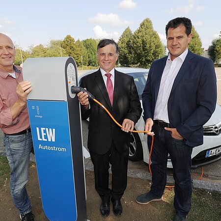 Drei Männer stehen neben einer LEW Autostrom-Ladestation mit dem Kabel in der Hand. Hinter Ihnen ein weißes Elektroauto auf einem Parkplatz. Im Hintergrund sind Bäume zu sehen.