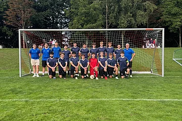 Fußballmannschaft mit 18 Spielern und 3 Trainern vor einem Tor auf Rasenplatz im Freien, Bäume im Hintergrund.