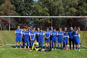 Jugendfußballmannschaft in blauen Trikots vor Tor auf grünem Spielfeld, im Hintergrund Bäume und zwei sitzende Personen.