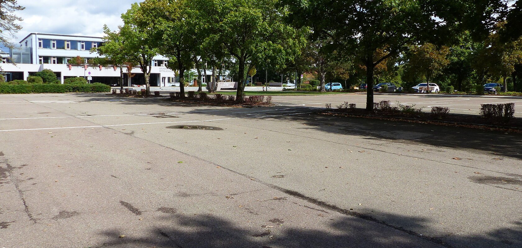 Großer leerer Parkplatz mit Bäumen und einem Gebäude mit Flachdach im Hintergrund unter blauem Himmel.