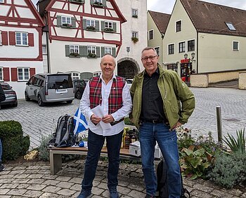 Zwei Männer stehen vor Fachwerkhäusern auf gepflastertem Platz und einer Bank mit Rucksack und Flaschen im Hintergrund. Die Bank ist umgeben von Pflanzen. Eine schottische Fahne - in weiß-blau - liegt auf der Sitzbank.