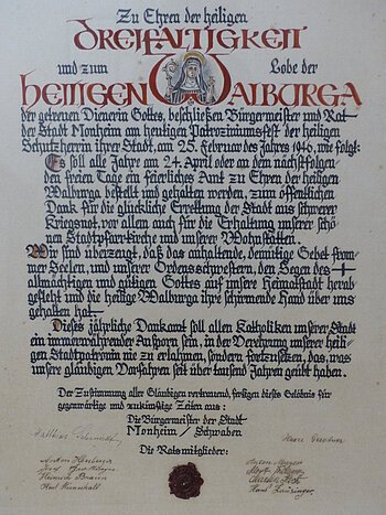 Ein handgeschriebenes Dokument mit dekorativen roten und schwarzen Buchstaben, das eine religiöse Widmung enthält. Hierin gelobt der Bürgermeister sowie der Rat der Stadt Monheim, ein alljährliches Dankamt zu Ehren der Heiligen Walburga abhalten zu lassen, für die Verschonung der Stadt im Zweiten Weltkrieg.