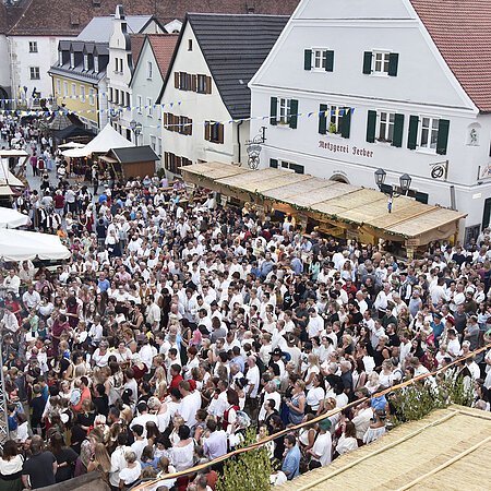 Große Menschenmenge auf dem historischen Stadtfest in der Altstadt von Monheim, mit Ständen und historischen Gebäuden im Hintergrund. Eine blau/weiße Flagge hängt an einem Gebäude.