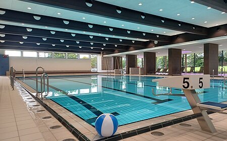 Innenaufnahme des leeren Schwimmbads - Jurabad Monheim. Zu sehen ist ein Schwimmbecken gefüllt mit strahlendem Wasser, ein Sprungbrett und ein weiß-blauer Wasserball liegt dekorativ im Vordergrund. Im Hintergrund des Hallenbads sind große Fenster zu sehen.