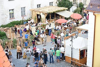 Menschenmenge teilweise in historischer Kleidung auf gepflastertem Platz vor einer dekorierten Holzhütte und einem historischen Gebäude dahinter, bei einer Veranstaltung im Freien. Der Platz um die Hütte ist mit Holzspännen gestreut und ist mit einem Holzzaun eingezäunt. Rechts im Bild weitere Stände, dabei vor einer weiß bedeckten Hütte ein WC-Schild an einer Straßenlaterne befestigt.