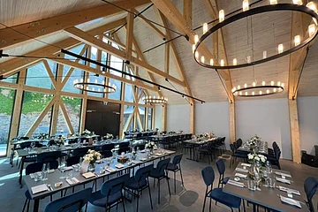 Großer Veranstaltungsraum einer Eventlocation mit offenem Dachstuhl und sichtbaren hellen Holzbalken oben an der Decke. In Reihen aufgestellte schwarze Tische mit denkelblauen Stühlen. Tische sind gedeckt mit Gläsern, Servietten, Besteck und Blumen in Vasen. Links im Bild große Glasfront ebenfalls mit hellen Balken. An der Decke große, runde, moderne Leuchten angebracht.