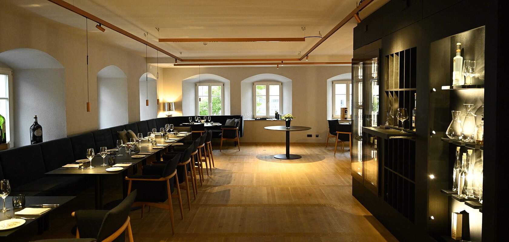 Innenansicht des Restaurants im Hotel by Ferber, in Monheim. Auf der rechten Seite ein großes schwarzes Regal mit verschiedenen Glasbehältern, Weingläsern und Flaschen. Auf der linken Seite schwarze Holztische mit schwarzen Sitzbänken und schwarzen Stühlen zu sehen.