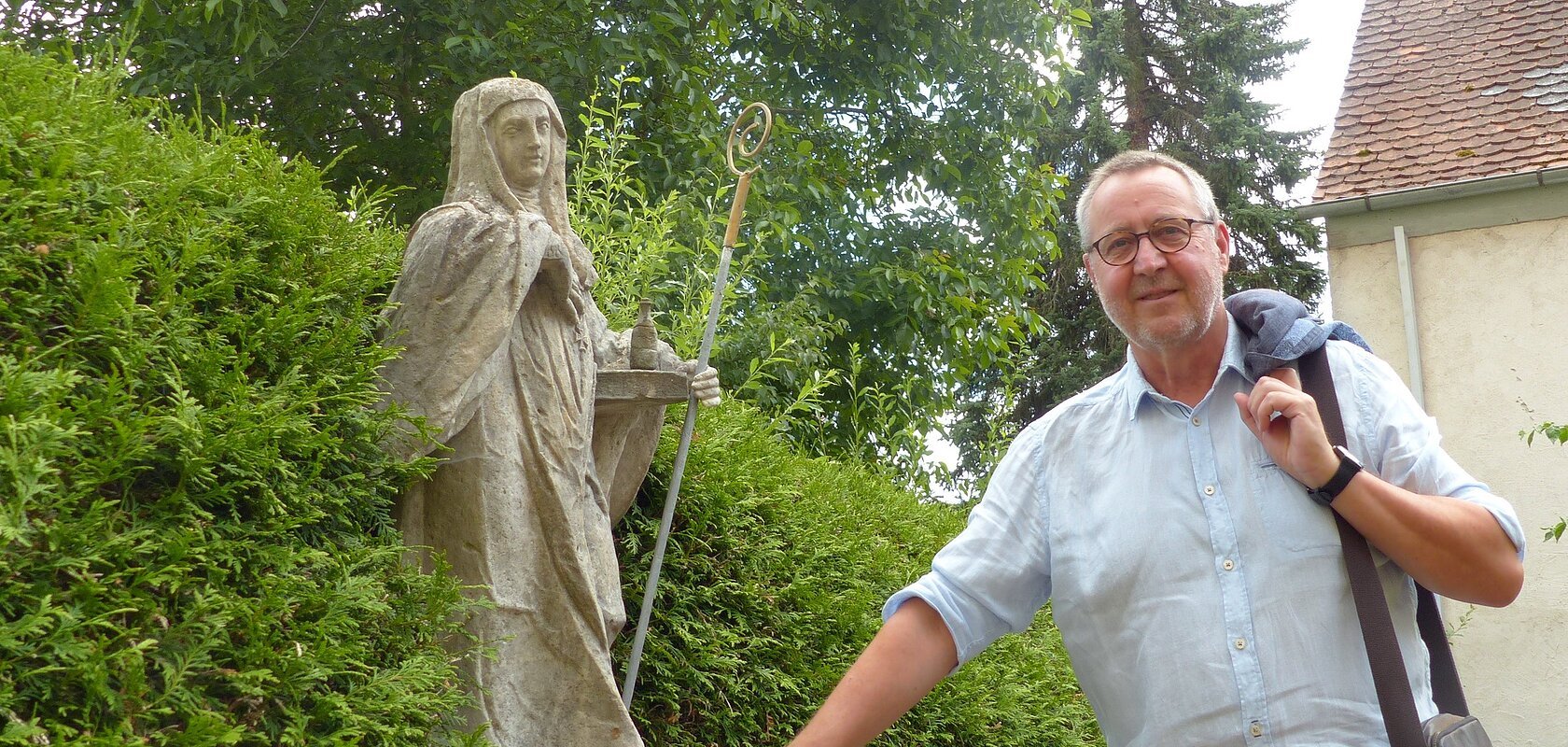 Eine Person mit Jacke über der Schulter steht neben einer Statue in einem Garten, umgeben von Büschen und einem Gebäude im Hintergrund.