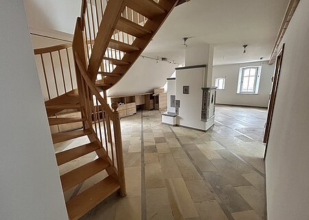 Mietobjekt Maisonetten Wohnung Marktplatz 11, Monheim.
Offener Raum mit Holztreppe, Fliesenboden, weißen Wänden und zwei Fenstern in einer Dachgeschosswohnung