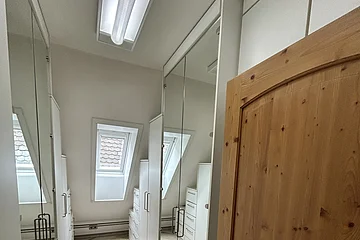 Miet-Maisonette - Ankleideraum. Schmaler Raum mit weißen Schränken, Spiegeln und einem Holz-Eingangstür, Dachfenster sichtbar.