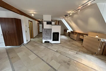 Miet Maisonette - Hauptraum - offener Bereich mit Kachelofen. Großer leerer Raum mit hellen Fliesen, Holzschränken, einem Kamin und schrägen Wänden unter einer Dachschräge.
