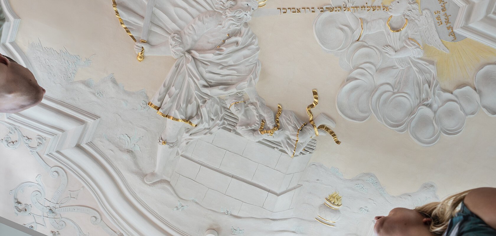 Zwei Personen betrachten ein kunstvoll verziertes Deckenrelief mit religiösem Motiv und hebräischer Schrift, die Stuckdecken im Rathaus Monheim.