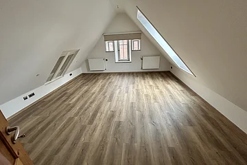 Leerer Raum mit schrägen Wänden, Holzfußboden, drei Fenstern und zwei Heizkörpern unter dem Fenster.