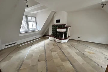 Miet Maisonette Marktplatz. Leerer Raum mit hellen Bodenfliesen, weiß gestrichenen Wänden, Dachfenster und grün-weißer Kachelofen in der Ecke.