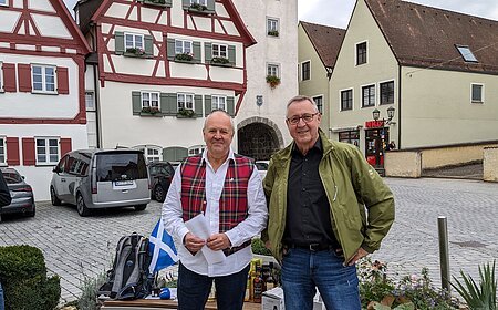 Zwei Männer stehen vor Fachwerkhäusern auf gepflastertem Platz und einer Bank mit Rucksack und Flaschen im Hintergrund. Die Bank ist umgeben von Pflanzen. Eine schottische Fahne - in weiß-blau - liegt auf der Sitzbank.
