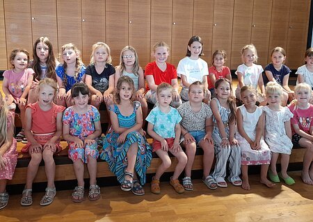 Zwei Reihen mit 22 Mädchen aus einem Kinderchor sitzen auf einer Holztreppe vor einer Holzpanelwand in einem Innenraum und blicken in die Kamera.