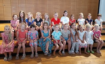 Zwei Reihen mit 22 Mädchen aus einem Kinderchor sitzen auf einer Holztreppe vor einer Holzpanelwand in einem Innenraum und blicken in die Kamera.