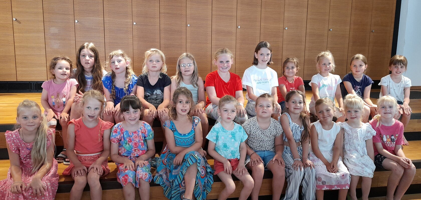 Zwei Reihen mit 22 Mädchen aus einem Kinderchor sitzen auf einer Holztreppe vor einer Holzpanelwand in einem Innenraum und blicken in die Kamera.