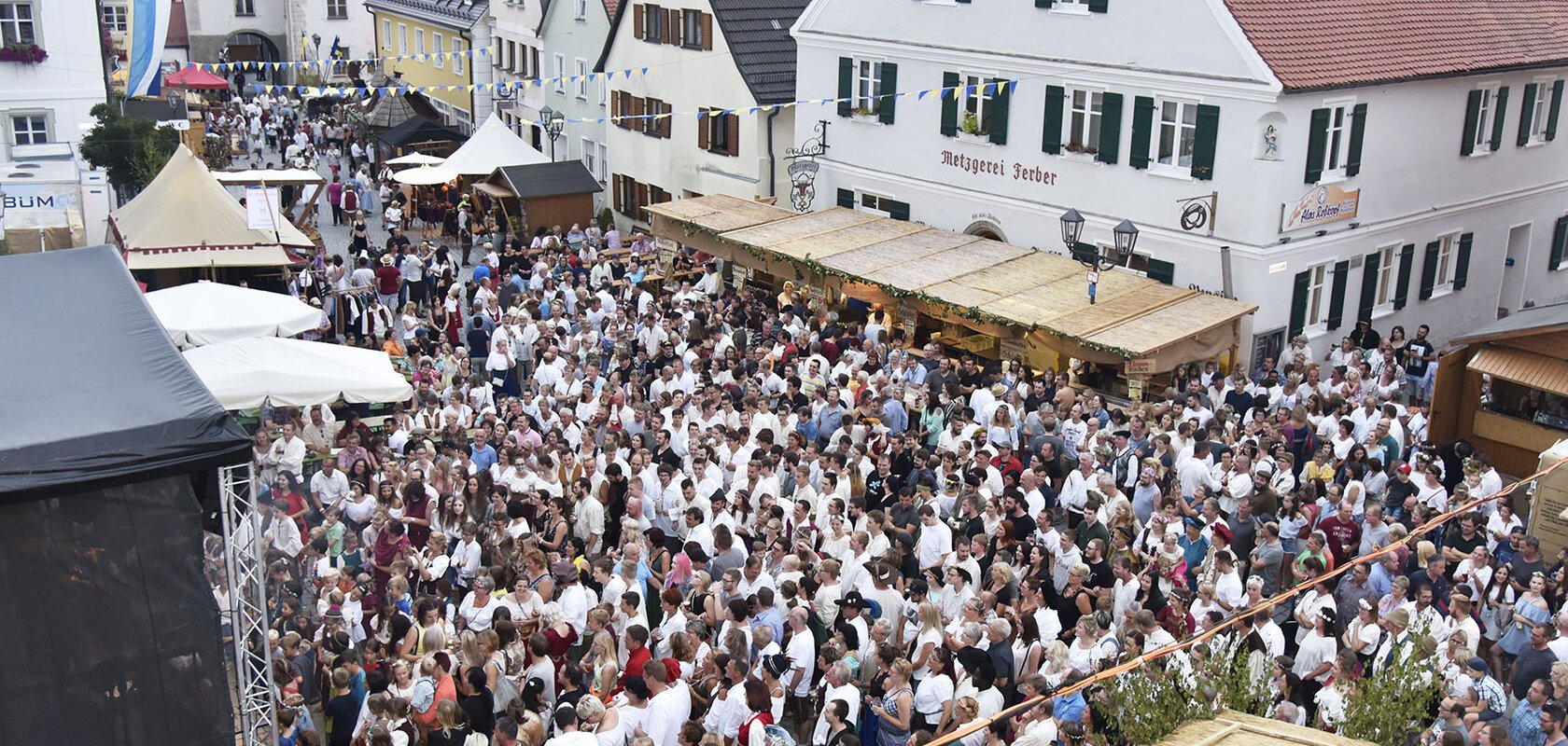 Große Menschenmenge auf dem historischen Stadtfest in der Altstadt von Monheim, mit Ständen und historischen Gebäuden im Hintergrund. Eine blau/weiße Flagge hängt an einem Gebäude.