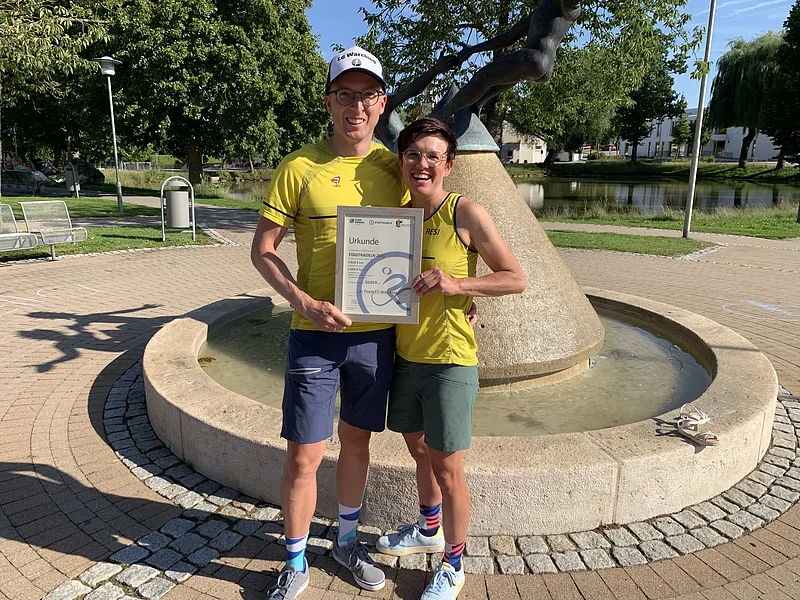 Zwei Personen in gelben Sportshirts und Shorts halten eine Urkunde vor einem runden Brunnen mit Skulptur, auf einem gepflasterten Platz, mit Bäumen und einem Weiher im Hintergrund.
