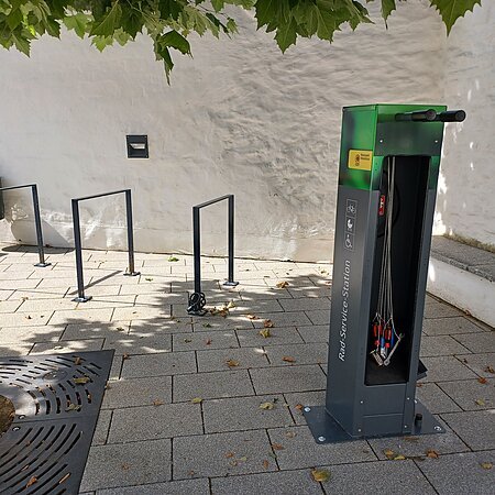 Fahrradservice-Station mit Werkzeugen und Fahrradständern auf einem gepflasterten Gehweg unter einem Baum. Umgeben von einer weißen Mauer.